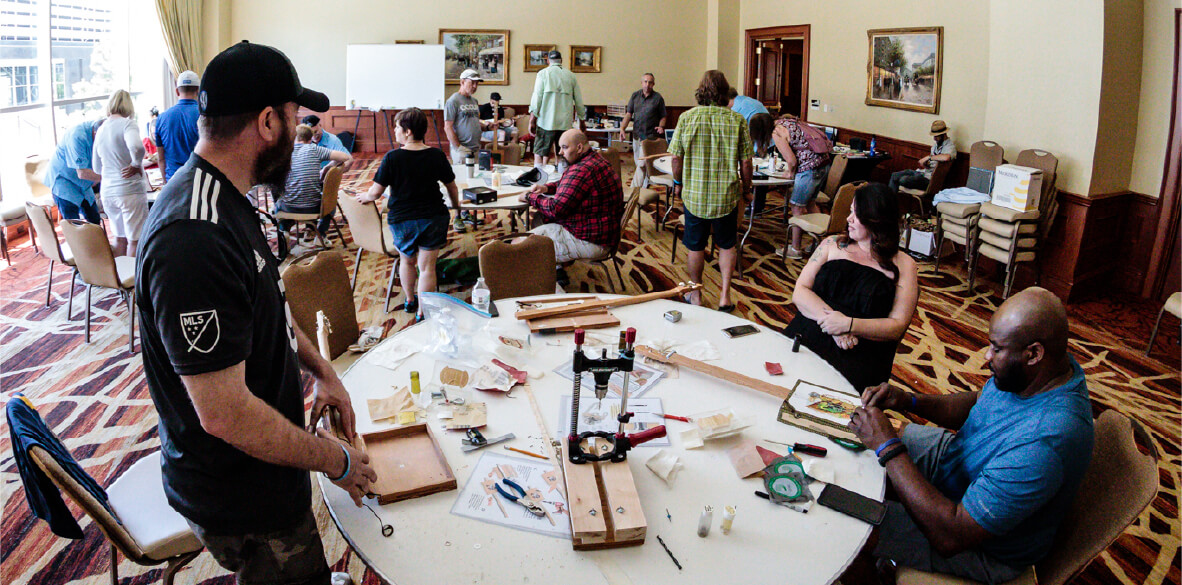 A lively group of people is engaged in a cigar box guitar-making workshop in a spacious room with large windows. The tables are cluttered with tools and guitar parts, as attendees focus on assembling their instruments. A man in the foreground is intently working on a guitar body, while others converse and continue their craft.