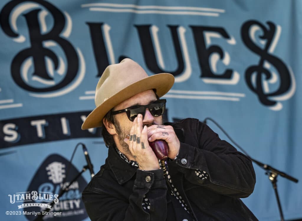 Blues musician Tony Holiday plays the harmonica on stage at the Utah Blues Festival, as indicated by the festival's logo on the blue banner in the background. He's wearing a tan fedora and black sunglasses, adding to his artistic stage presence. There is a watermark in the lower left indicating the image is from the Utah Blues Festival, and is Copyright 2023 by Marilyn Stringer.