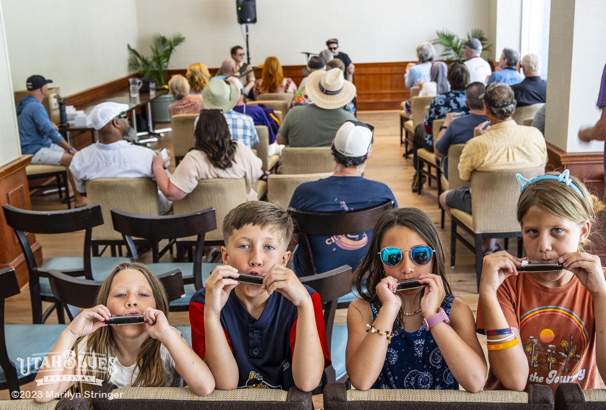 Four children are in the foreground at a harmonica workshop, each holding a harmonica to their mouths, with looks of concentration. They are seated inside a well-lit room with a crowd of adults behind them, attentively watching a musician conducting the workshop on a small stage. There is a watermark in the lower left indicating the image is from the Utah Blues Festival, and is Copyright 2023 by Marilyn Stringer.