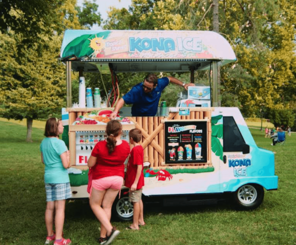 A Kona Ice truck serving shaved ice to children in a park, with a colorful tropical design and flavor station on the side.