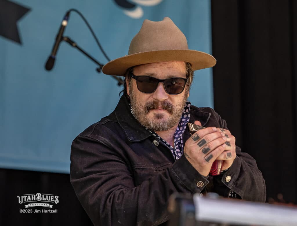 Tony Holiday performing at the Utah Blues Festival, wearing a tan hat, sunglasses, and dark jacket while holding a harmonica.