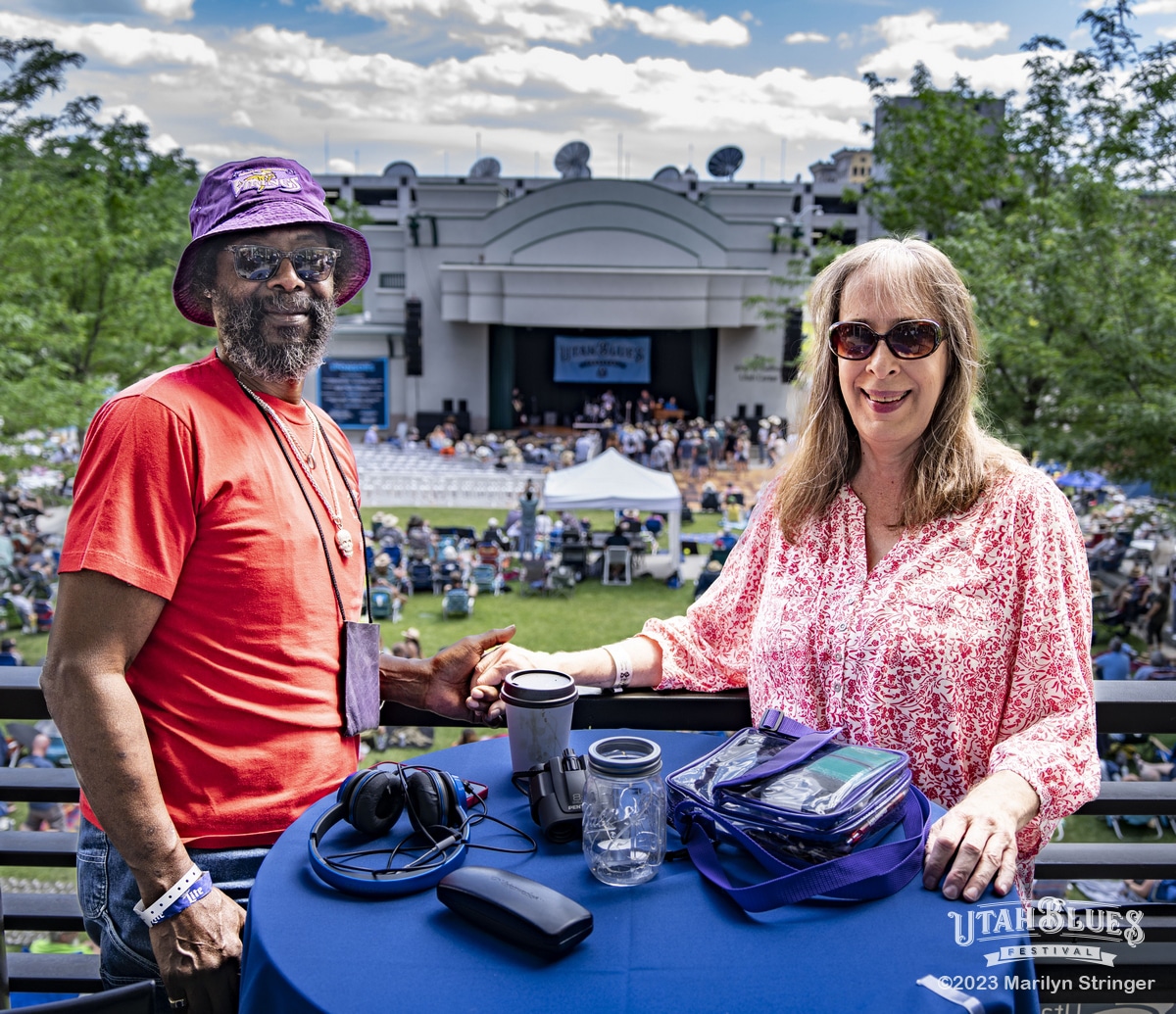 Two people smile at an outdoor music festival while standing at a balcony table overlooking the stage and crowd. The man wears sunglasses, a purple bucket hat, and a red shirt, while the woman wears sunglasses and a pink patterned blouse. They hold hands across the table, which has a coffee cup, headphones, and other items on it. The stage and audience of the Utah Blues Festival are visible in the background under a partly cloudy sky.