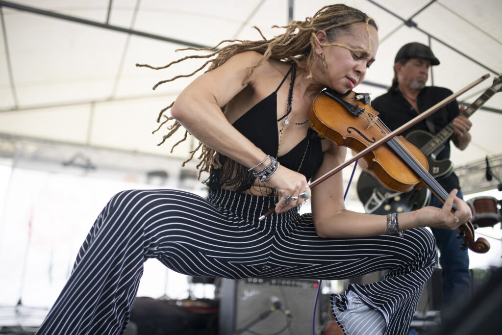 Anne Harris performing energetically onstage, playing a violin while wearing a black fringe top and striped pants, with long braided hair flowing as a guitarist performs in the background.