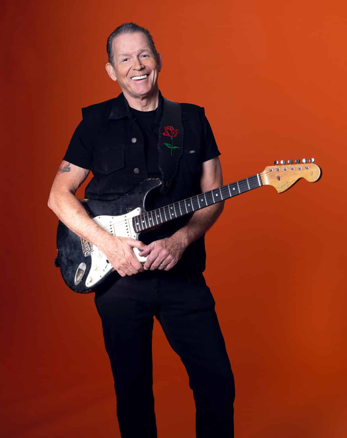 Tommy Castro smiling while holding a worn black electric guitar, dressed in black against a vibrant orange studio backdrop.