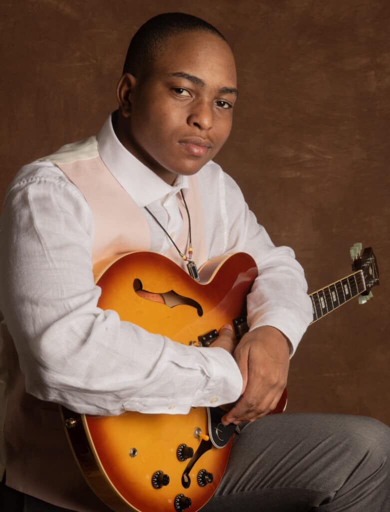 Harrell “Young Rell” Davenport seated with a sunburst electric guitar resting upright against his chest, wearing a peach vest and white shirt in front of a brown backdrop.