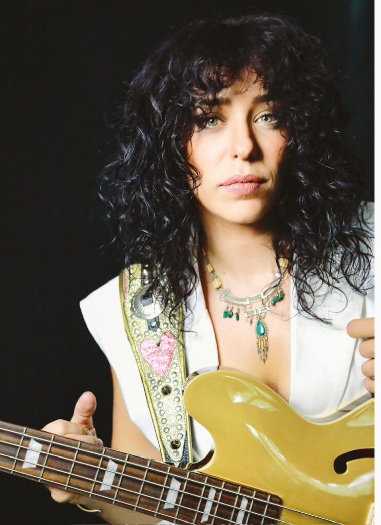Danielle Nicole, a musician with curly dark hair holding a gold electric bass guitar, wearing layered necklaces and a white top, with a custom strap featuring a pink heart design against a dark background.