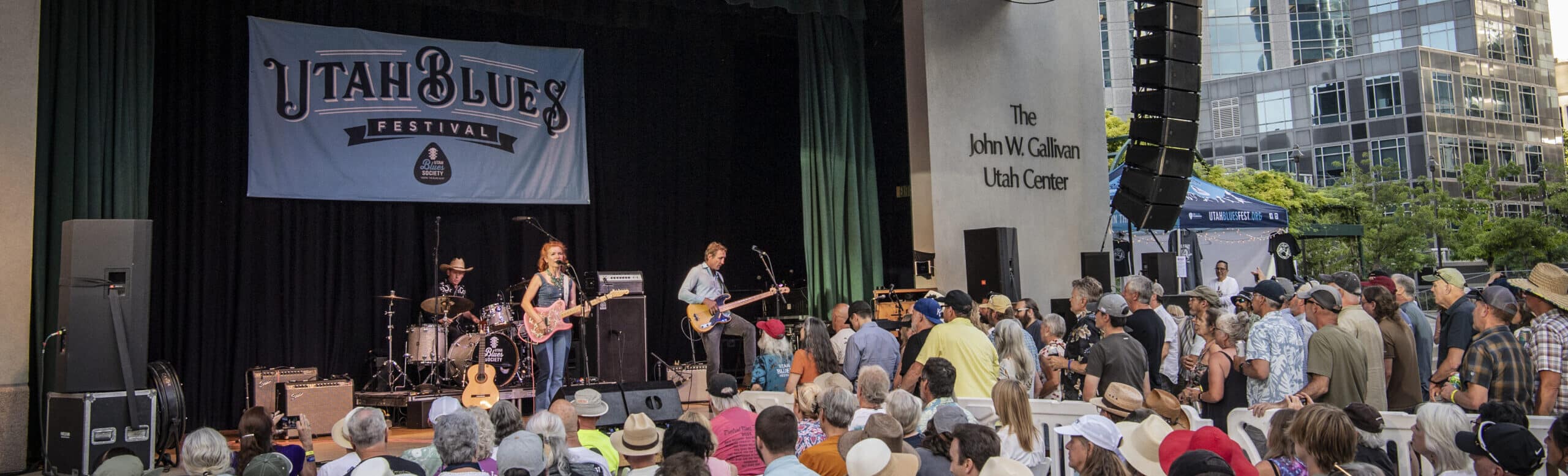 Utah Blues Festival live performance at the Gallivan Center, with a band playing on stage to a seated outdoor audience in downtown Salt Lake City.